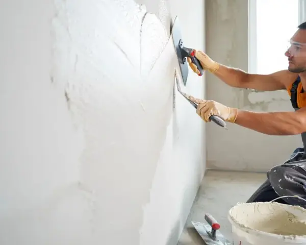 Un ouvrier applique un enduit sur un mur humide avec une truelle et une taloche. Le travail progresse, laissant une surface lisse.
