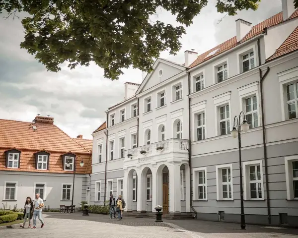 Elegancki pałac Zwartowo z białymi zdobieniami i łukowym wejściem. Na pierwszym planie para spaceruje po brukowanym dziedzińcu.
