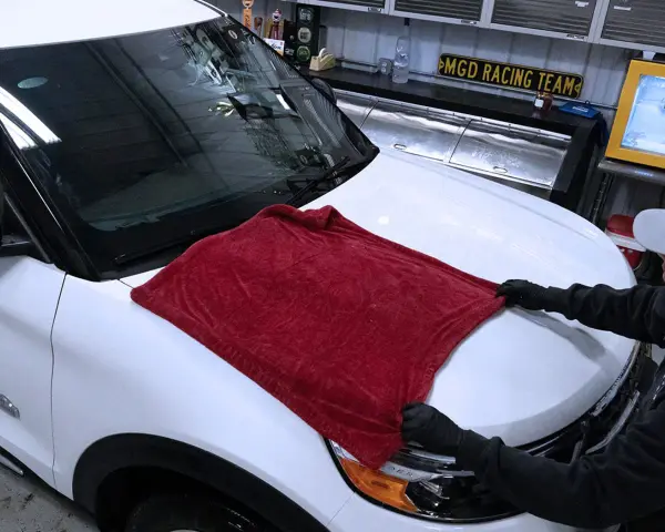 Person in gloves carefully places a plush red towel on a white car hood, demonstrating the best way to wash a car without scratching.