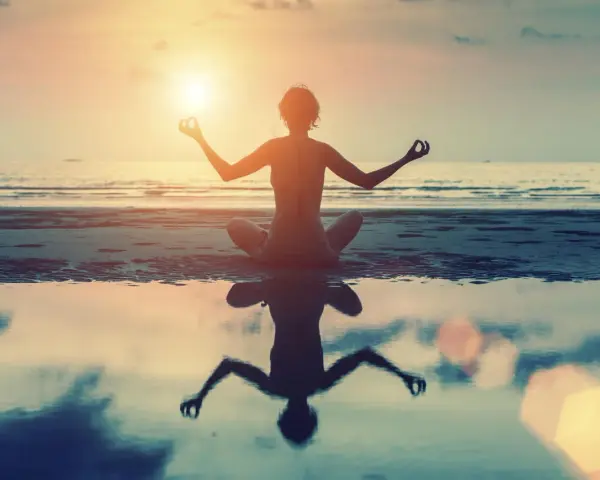 Frau in Lotusposition am Strand bei Sonnenuntergang. Ihre Meditation stärkt das Selbstbewusstsein.