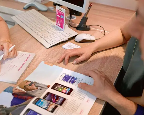 Dwie osoby przeglądają ofertę telefonów komórkowych. Na biurku leży klawiatura, myszka, skaner i katalog z produktami. Jedna z osób pisze coś na formularzu.