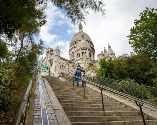 Dwie osoby wspinają się po schodach na wzgórze Montmartre, w tle Bazylika Sacré-Cœur.