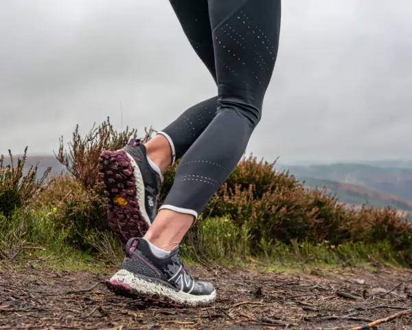 Gambe di una donna che fa trail run su un sentiero fangoso, con erica e colline nebbiose sullo sfondo.