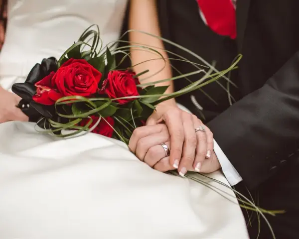 Dłonie nowożeńców splecione na tle bukietu czerwonych róż. Idealny moment, by pomyśleć, jak przygotować życzenia ślubne które zapadną w pamięć i wzruszą.