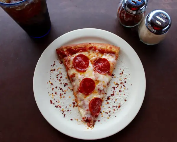 Kawałek pizzy pepperoni na talerzu, posypany płatkami chili i parmezanem. Idealny na szybki lunch, choć warto pamiętać o jego kcal.
