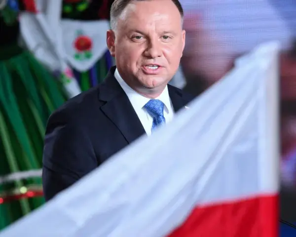 Prezydent Andrzej Duda w garniturze i krawacie, na tle polskiej flagi i tradycyjnego stroju. Ile trwa ułaskawienie prezydenta?