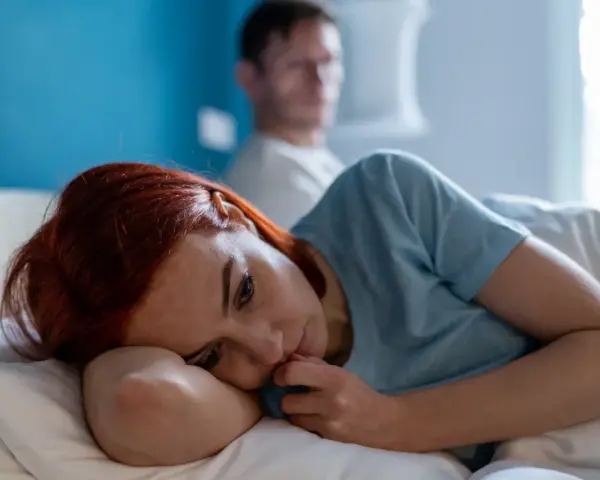 Zamyślona kobieta z rudymi włosami leży w łóżku, zastanawiając się: "czy jestem toksyczna?". W tle niewyraźny mężczyzna.