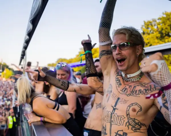 Feiernde Menschenmenge bei einem Festival, die zu **techno music** tanzt. Ein Mann mit Tattoos und Sonnenbrille jubelt.