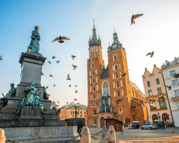 A monument and St. Mary's Basilica dominate this Krakow travel scene. Birds fly around the historic architecture under a clear sky.