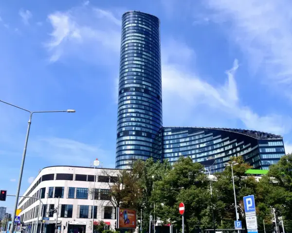 Wysoki, szklany budynek Sky Tower we Wrocławiu, z pewnością ma wiele pięter. Widać też niższy budynek i drzewa.