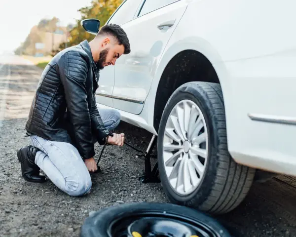 Mężczyzna klęczy przy białym samochodzie, używając podnośnika, by podnieść auto bez kanału. Obok leży zapasowa opona.