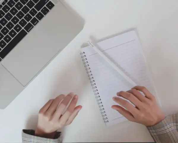 Ręce piszą CV po niemiecku przy laptopie, notatniku i kawie.
