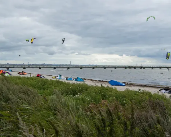 Kitesurfare förbereder sig vid Habo Ljung Camping. Många kites svävar i luften över en strand med en lång pir i bakgrunden.