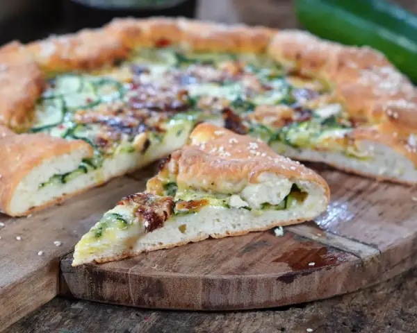 Leckeres Zucchini Pizza Rezept auf einem Holzbrett, mit einem Stück abgetrennt.