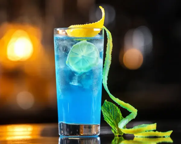 Niebieski koktajl z granatem, lodem, plasterkiem limonki i ozdobną skórką cytrynową.