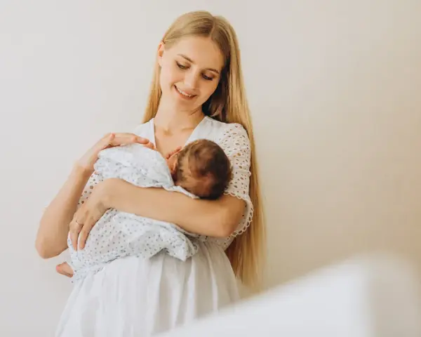 Uśmiechnięta młoda mama tuli noworodka, karmiąc go piersią. Delikatne spojrzenie i biała sukienka podkreślają jej macierzyństwo.