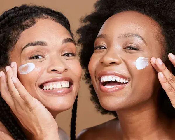 Deux femmes souriantes appliquent une crème sur leur visage, un geste pour une exfoliation visage réussie.
