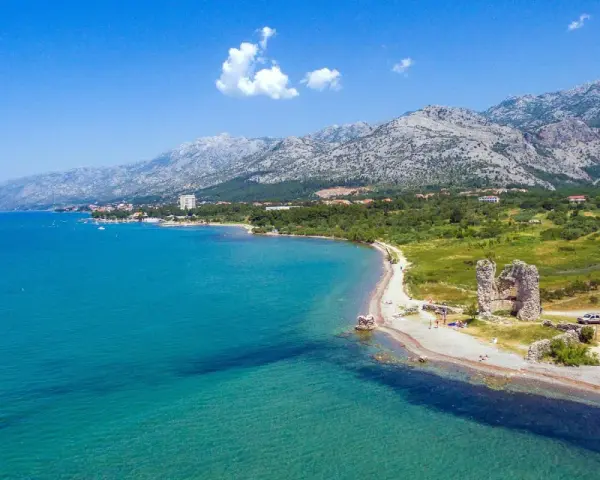 Piękne wybrzeże z górami w tle, idealne na camping Paklenica. Woda kusi turkusowym kolorem, a na plaży widać ruiny starej twierdzy.