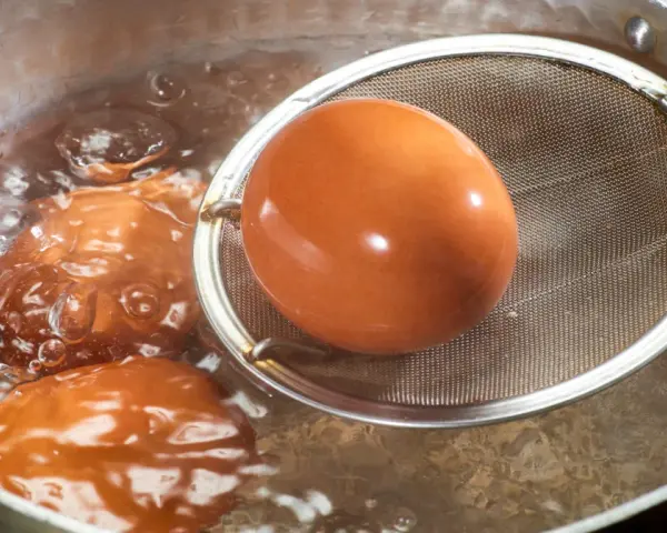 Jak ugotować jajka? Brązowe jajka gotują się w bulgoczącej wodzie, jedno z nich jest wyjmowane sitkiem.