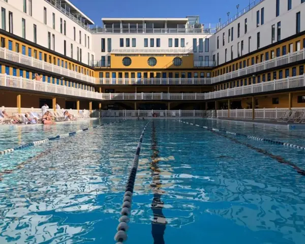 Piscine art déco parisienne, avec ses bâtiments jaunes et blancs, ses balcons et ses transats. L'eau bleue invite à la baignade.