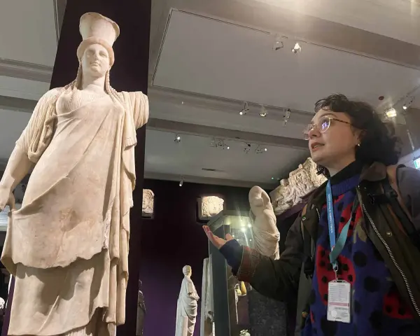 A woman with a museum pass for Turkey gestures towards a marble statue of a woman in a museum.