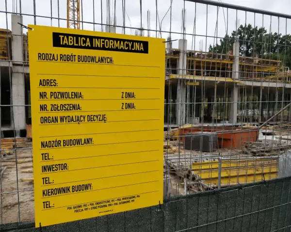 Żółta tablica informacyjna budowy z danymi o pozwoleniu, adresie i kontaktach. W tle widać konstrukcję budynku.