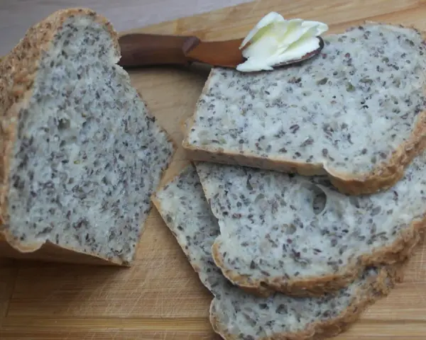 Chleb na proszku do pieczenia, bogaty w ziarna, pokrojony na kromki. Na jednej z nich leży masło na drewnianej łopatce.