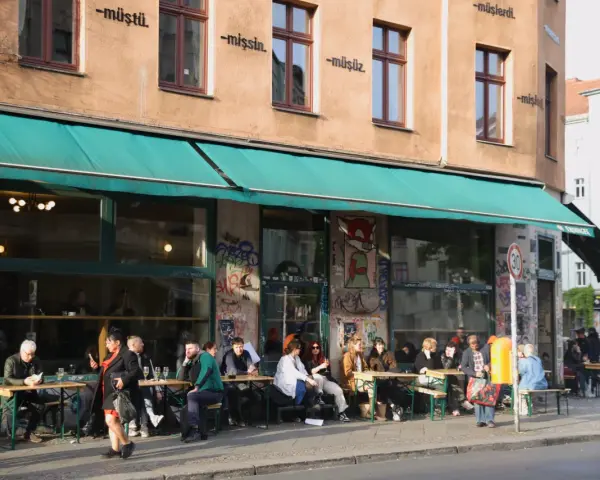 Menschen sitzen an Tischen vor einem Café in Berlin. Die Fassade ist mit Graffiti verziert und trägt die Aufschrift "dorle berlin".