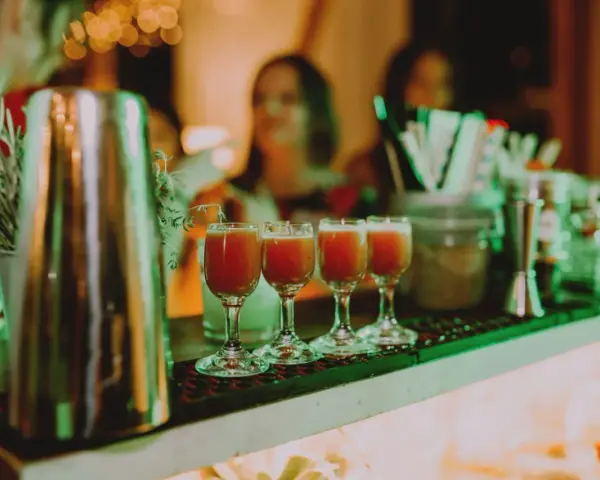 Cztery kieliszki z czerwonym drinkiem stoją na barze, gotowe do podania na weselu. W tle widać gości i dekoracje.