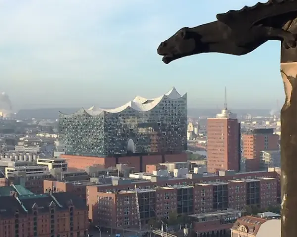 Blick über Hamburg mit der Elbphilharmonie, gesehen durch ein Fenster des Mahnmals St. Nikolai. Ein Wasserspeier wacht über die Stadt.