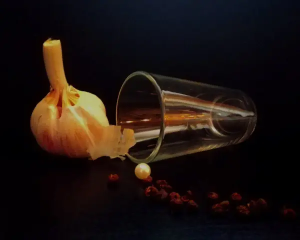 Photographie nature morte : une tête d'ail, un verre renversé, une perle et des baies de poivre rouge sur fond sombre.