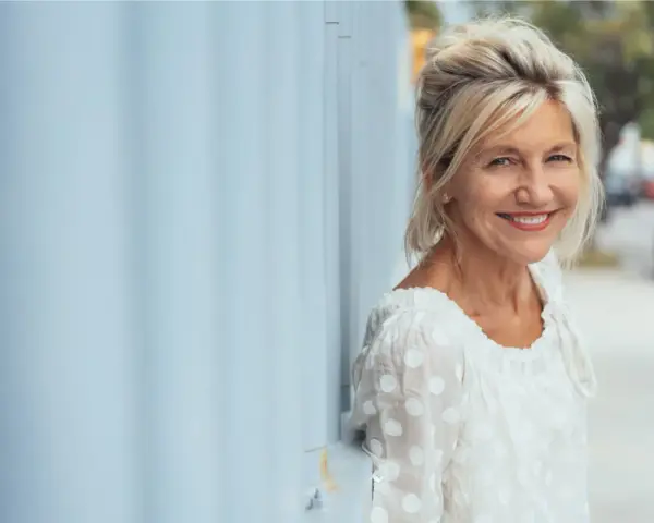 Donna sorridente appoggiata a un muro blu. Potrebbe beneficiare di acido folico in menopausa per il suo benessere.