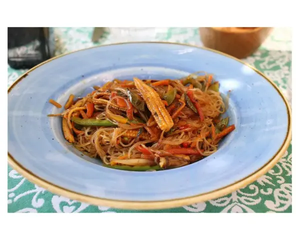 Un piatto di spaghetti di riso con verdure colorate, tra cui peperoni, carote e pannocchiette, condito con una salsa saporita.