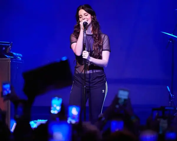 Lana Del Rey, w czarnej, prześwitującej bluzce i sportowych spodniach, śpiewa na scenie. Fani z telefonami tworzą tłum. To jej lana del rey opener.