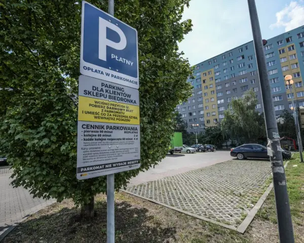 Płatny parking dla klientów Biedronki. Obok drzewa stoi znak informujący o cenniku i zasadach parkowania.