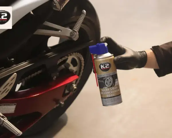 Ręka w czarnej rękawiczce aplikuje suchy smar do łańcucha motocyklowego K2. Idealny wybór, by zadbać o napęd.