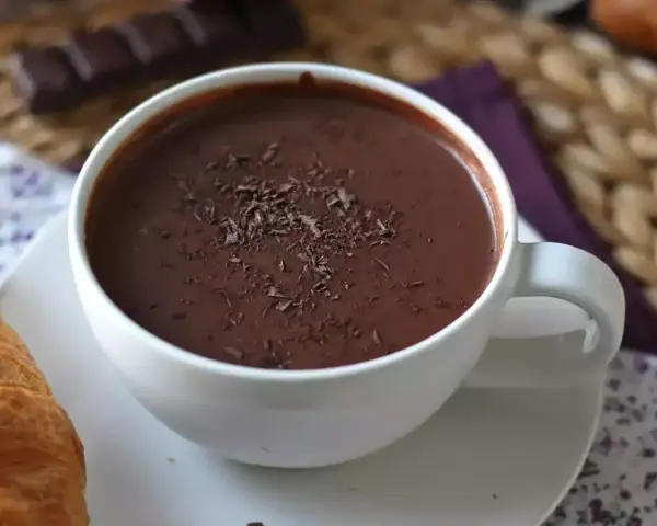 En vit mugg med varm choklad, toppad med chokladspån. En croissant ligger bredvid.