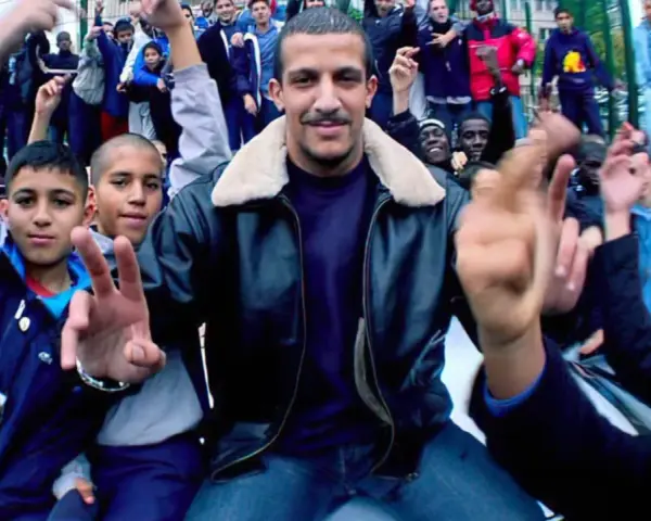 Un groupe de jeunes pose pour une photo, certains font des signes de paix. L'ambiance est celle du rap francais.