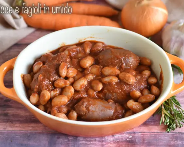 Piatto fumante di salsiccia e fagioli in umido, con carote, cipolla e aglio sullo sfondo.