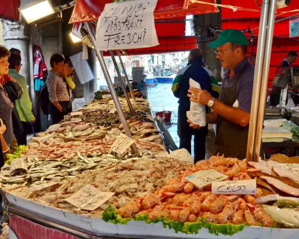 Mercato Venezia: bancarella di pesce fresco con gamberi, calamari e pesciolini. Venditore con cappellino verde.