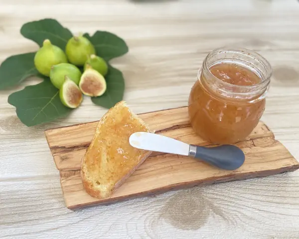 Fetta di pane con marmellata di fichi, accanto a un vasetto e a fichi freschi su un tagliere di legno.