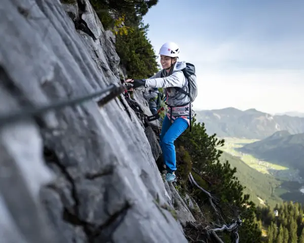 Kobieta w kasku i uprzęży pokonuje via ferrata, chwytając się stalowej liny. W tle rozciąga się malownicza górska dolina.