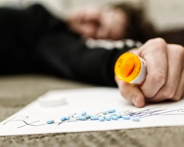 Dłoń trzymająca otwartą butelkę z lekami, obok rozsypane tabletki na kartce z napisem "Rx".