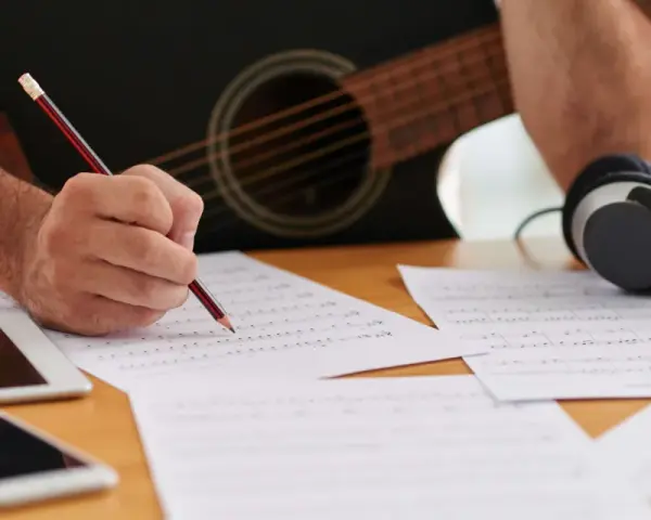 En person skriver noter med en penna, med en gitarr och hörlurar i bakgrunden. Detta är en bra inledning till en ny låt.
