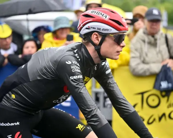 Trine Vingegaard, a cyclist in a grey and black racing suit, rides in the rain. Spectators watch from behind a yellow barrier with "Tour de France" branding.