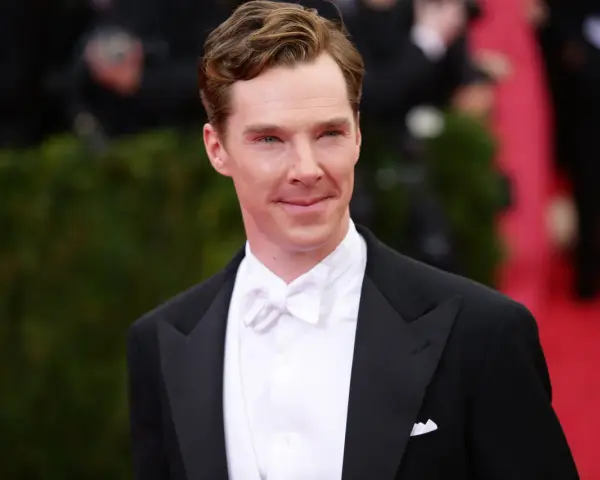 Benedict Cumberbatch w czarnym smokingu i eleganckim białym krawacie, z uśmiechem pozuje na czerwonym dywanie.
