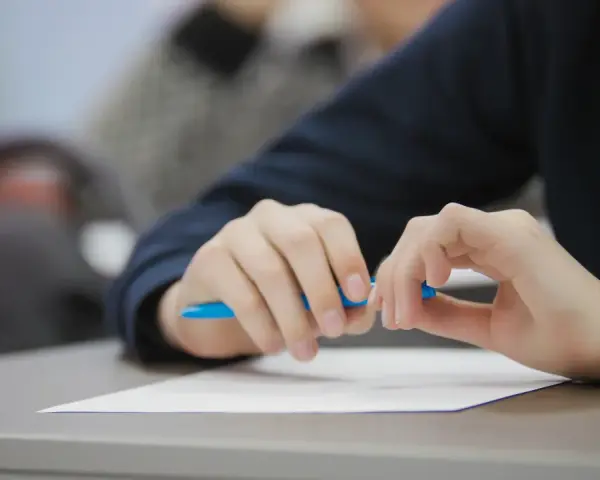 Uczeń pisze wypracowanie, przygotowując się do matury 2026. Na biurku biała kartka i niebieski długopis. Lektury obowiązkowe czekają.