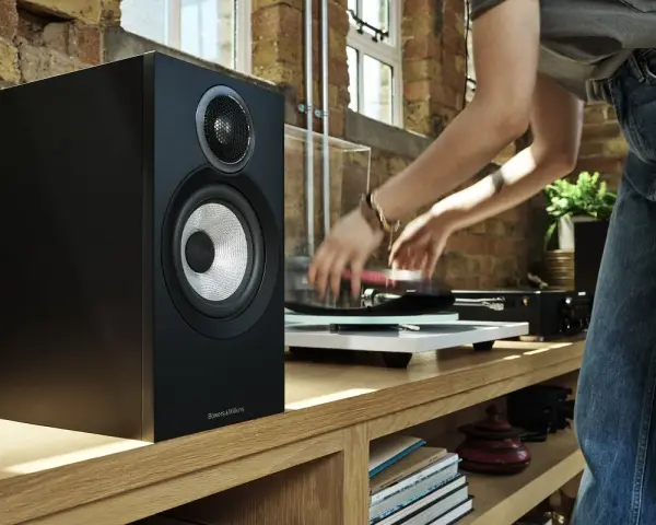 Czarny głośnik Bowers & Wilkins, jeden z najlepszych producentów głośników, obok gramofonu, na którym osoba kładzie płytę.