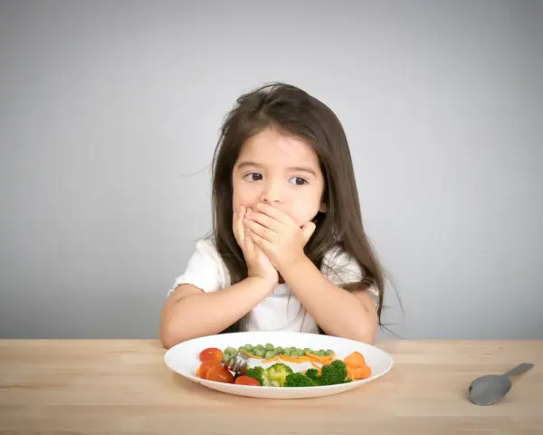 Kleines Mädchen bedeckt Mund, schaut auf Teller mit Gemüse. Manche kinder essen schnell, aber dieses hier scheint zu überlegen.