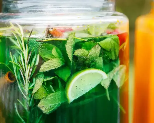 Zielony, orzeźwiający syrop z mięty z dodatkiem limonki i rozmarynu w szklanym słoju. Obok pomarańczowy napój.